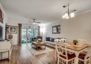 A spacious living room with a wooden floor, a dining table with white chairs, a sofa, and a ceiling fan. - New on Program Cypress Bend 1124! Updated and Close to Beach and Golf! (North Myrtle Beach)