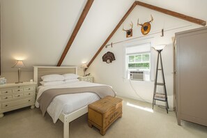Ein Schlafzimmer mit geneigter Decke, einem Bett mit weißer Bettwäsche, einer Holzkiste, einem Nachttisch, einer Lampe, einer Leiter und Wandleuchtern aus Hirschgeweihen.
