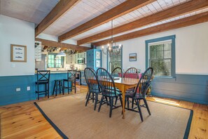 A dining area with a wooden table and chairs, a kitchen in the background, and a view through a window. - Heron's Perch Beautiful Pondside Home with Screened Porch (Brunswick)