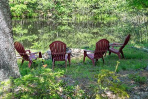 Three Adirondack chairs facing a calm lake surrounded by greenery. - Heron's Perch Beautiful Pondside Home with Screened Porch (Brunswick)