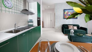 A modern kitchen with green cabinets, a white countertop, and a clock. There is a dining area with a table set for two, a sofa, and a decorative painting on the wall. - Estate4home - Velia in Sorrento (Sorrento)
