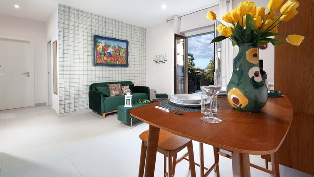 A modern dining area with a wooden table, chairs, and a vase of yellow tulips. There is a green sofa and a painting on the wall. - Estate4home - Velia in Sorrento (Sorrento)