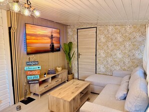 Ein Wohnzimmer mit einem TV-Schrank aus Holz, einer Couch und einem Couchtisch. An der Wand hängt ein Gemälde von einem Sonnenuntergang, und eine Tür ist mit ornamentalem Wandpapier versehen.