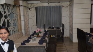 A restaurant with a long dining table set for a meal, a waiter standing in front of it, and a decorative wall with a curtain in the background. - HOTEL  RAJ PALACE SHIVPURI (Shivpuri)
