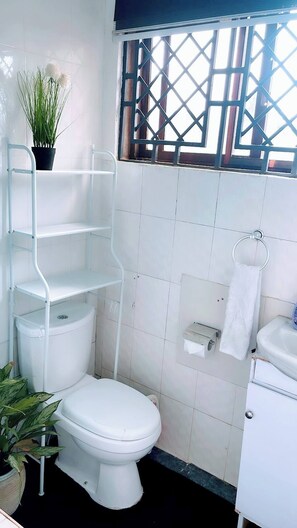 A bathroom with a toilet, a sink, a toilet paper holder, and a window with a lattice design. - Cozy Studio Apartment in Vibrant Spintex, Accra (Accra)