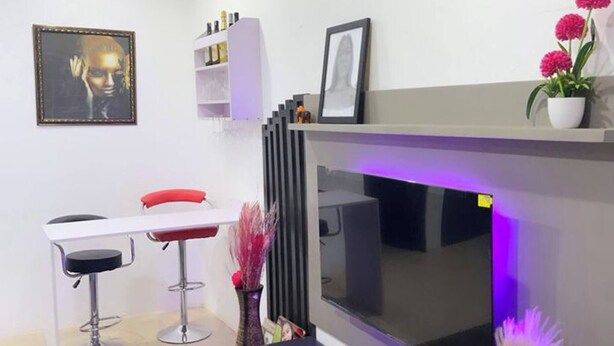 A modern living room with a large flat-screen TV, a coffee table, two bar stools, a white shelf with decorative items, and a framed picture on the wall. - Cozy Studio Apartment in Vibrant Spintex, Accra (Accra)
