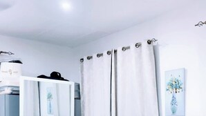A bedroom with a bed, a grey tufted headboard, white curtains, and a small bedside table. - Cozy Studio Apartment in Vibrant Spintex, Accra (Accra)