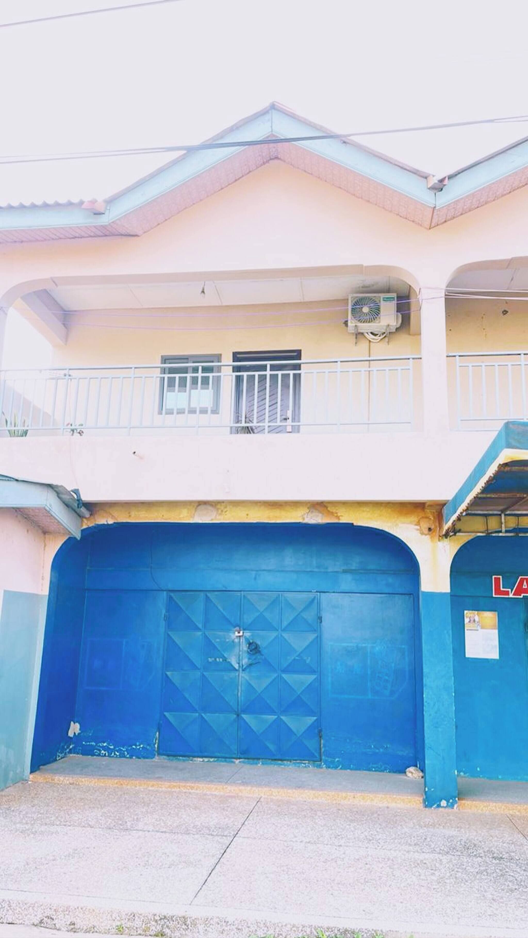 A two-story building with a blue garage door, a balcony with a railing, and a small air conditioning unit mounted on the wall.