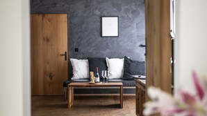 Ein modernes Wohnzimmer mit Holzboden, einer dunkelgrauen Wand, einem schwarzen Sofa mit weißen Kissen, einem Holztisch und einem gerahmten Bild über dem Sofa.