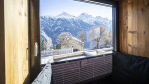Ein Interieur einer Hütte mit einem großen Fenster, das einen Blick auf schneebedeckte Berge und ein kleines Gebäude mit weißem Zaun bietet.