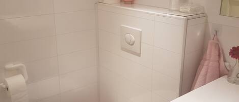 A small, white bathroom with a toilet, a towel rack with a pink towel, and a glass container on the sink.