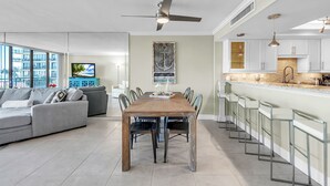 Una sala de estar moderna con comedor, cocina y un amplio ventanal con vistas a un paisaje urbano.