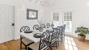 A dining room with a white table set for six, surrounded by black chairs with woven seats. A large chandelier hangs from the ceiling, and a mirror with a black frame is mounted on the wall. - "One Particular Harbor" BHI and Shoals Club, 5 BR 3.5 BA, New Rental 2026 (Bald Head Island)