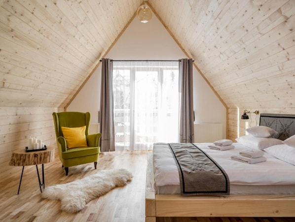 A room with a sloped ceiling, a bed with white linens, a green armchair, a small round table with candles, and a fur rug.