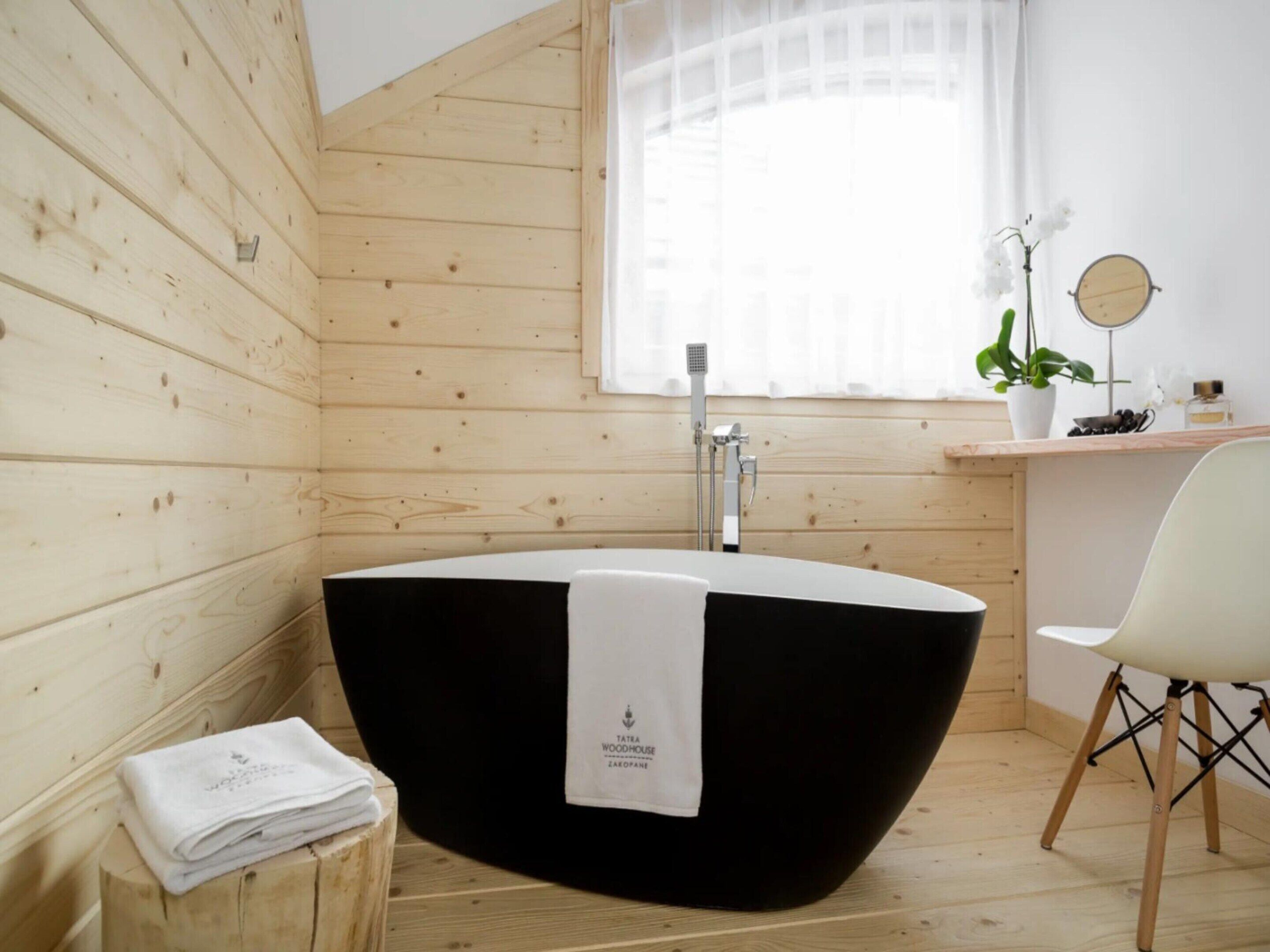 A modern bathroom with a freestanding bathtub, wooden walls, a window with sheer curtains, a small mirror, and a potted plant.