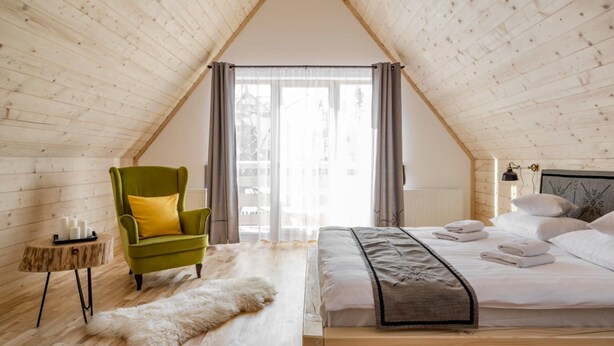 A cozy bedroom with a sloped ceiling, a bed with neatly folded towels, a green armchair, a fluffy rug, and a small side table with candles. - Comfortable Holiday Home, 3 Bedrooms, Fireplace (Zakopane)