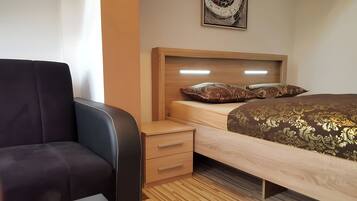 Um quarto com uma cama, uma mesa de cabeceira de madeira com gavetas, uma cadeira preta e um ponto de luz fixado na parede.