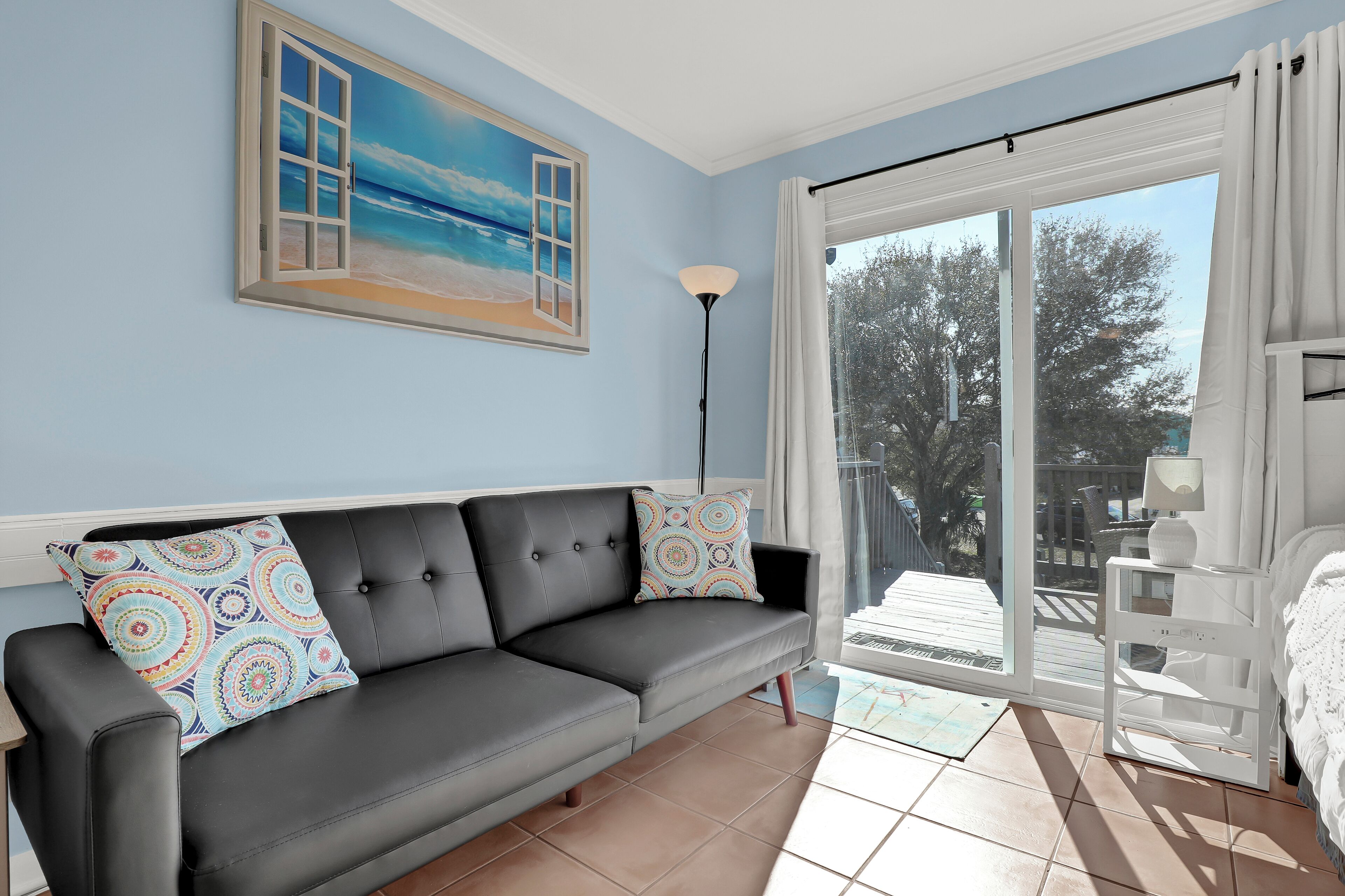 A room with a black leather sofa, a colorful pillow, a painting of a beach, a floor lamp, and a view through a sliding glass door.