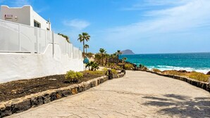 Ein gepflasterter Weg führt zu einem Strand mit Palmen und einem weißen Gebäude mit einem Schild.