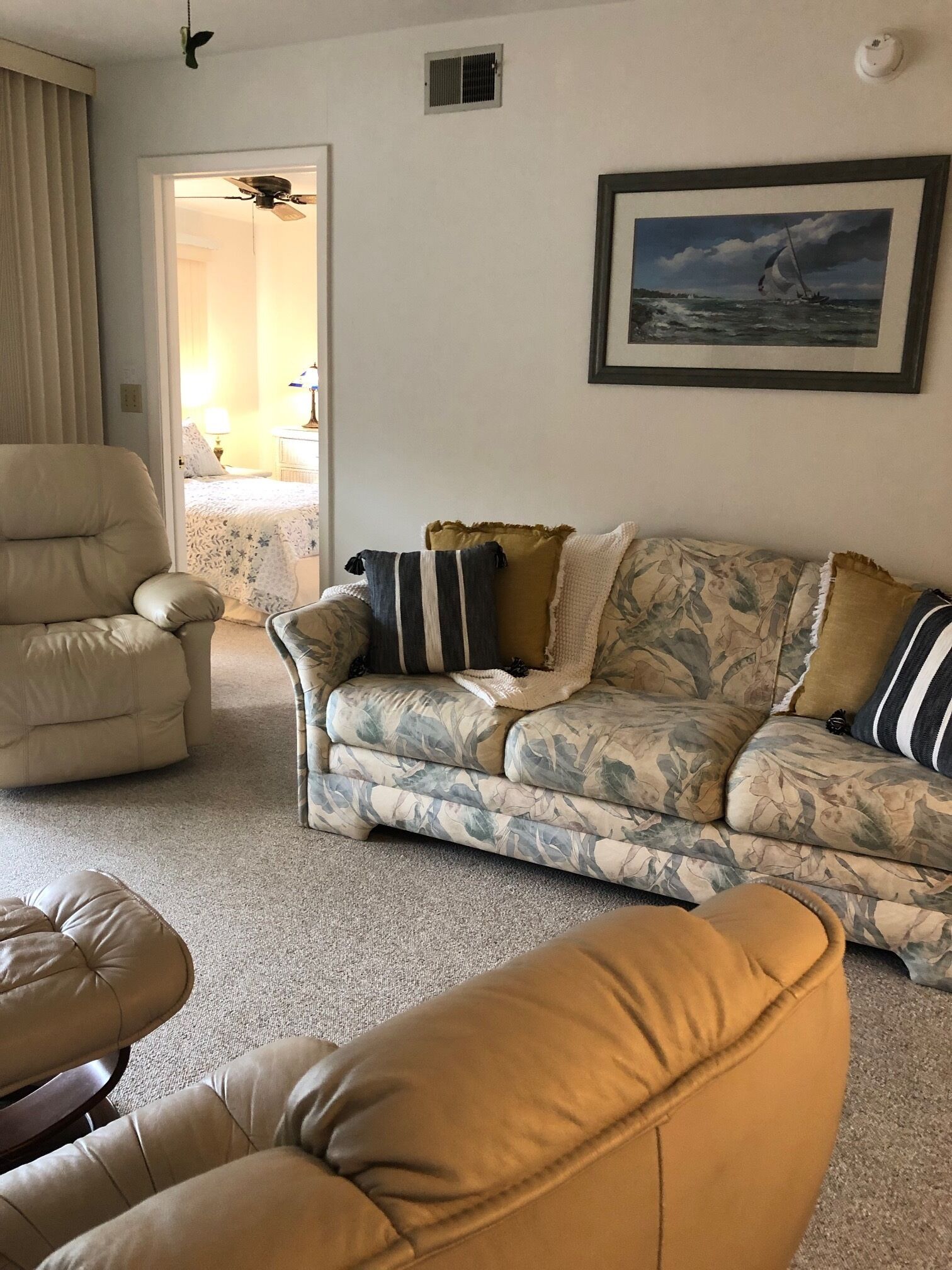 Una sala de estar con un sillón color beige, un sofá estampado con motivos florales y un reposapiés color arena. Se observa un ventilador de techo, un cuadro enmarcado en la pared y una puerta que da a otra habitación.