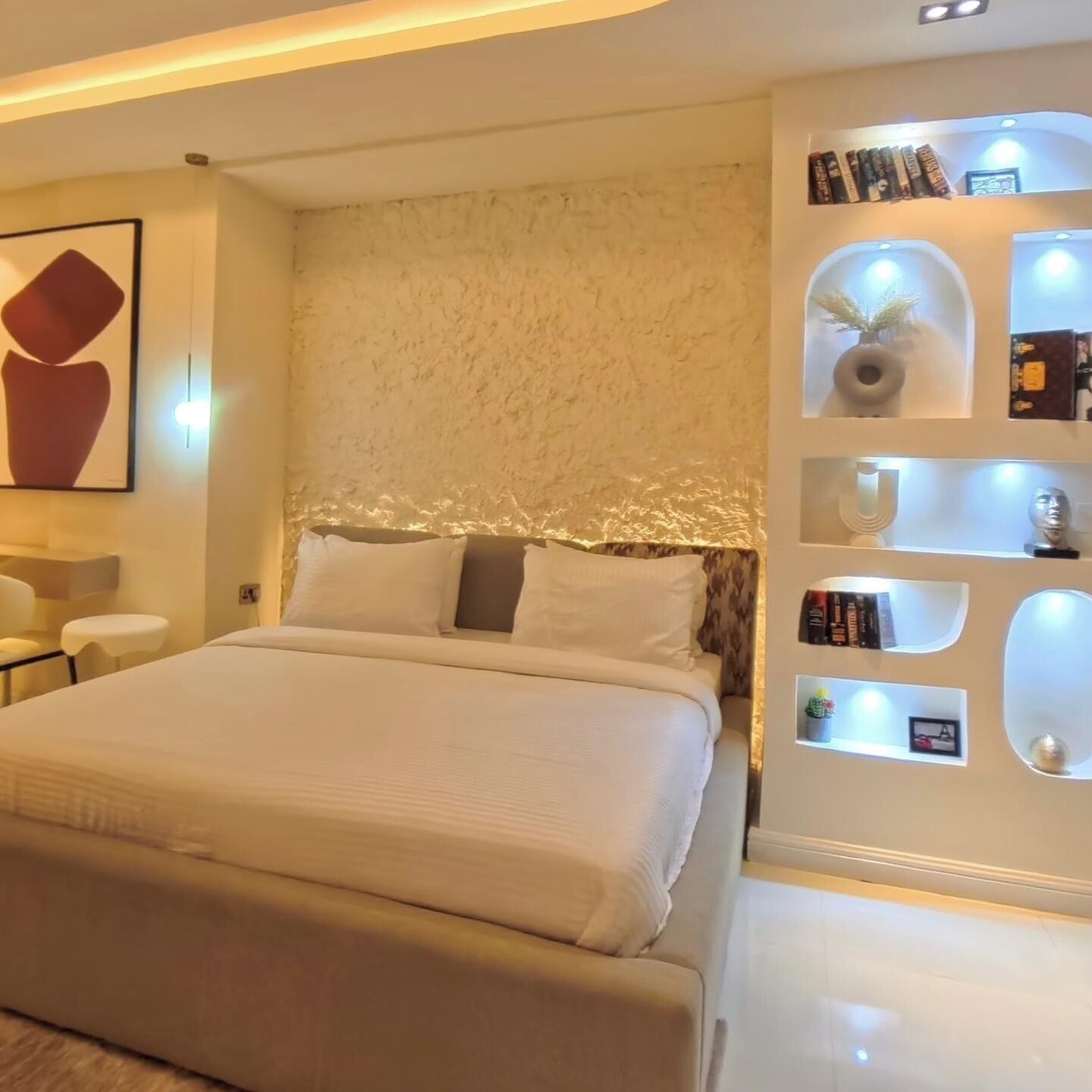 A modern bedroom with a large bed, a wall-mounted shelf with books and decorative items, and a minimalist design.