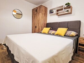 Een slaapkamer met een groot bed, een houten kledingkast, een nachtkastje en een wandplank met boeken en een plant.