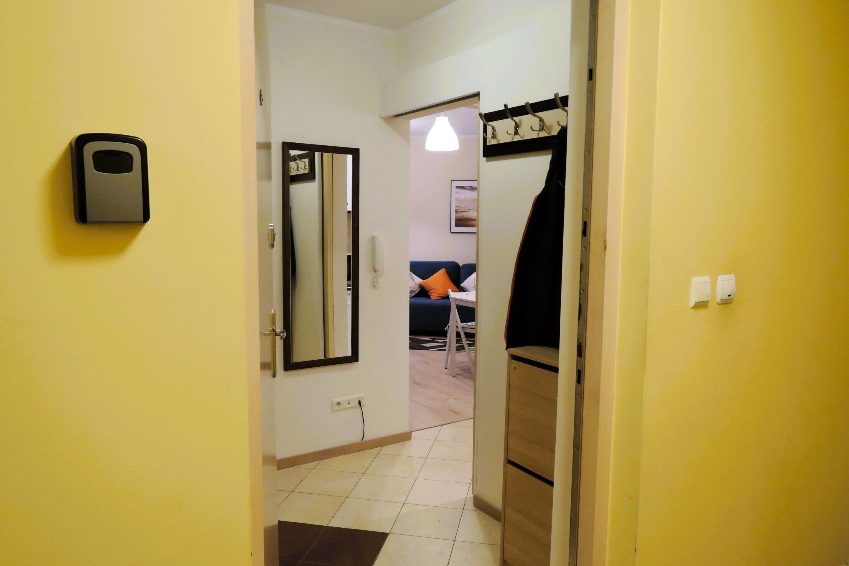 Un couloir avec un miroir, un porte-manteau et une porte donnant sur un salon équipé d’un canapé et d’une lampe.