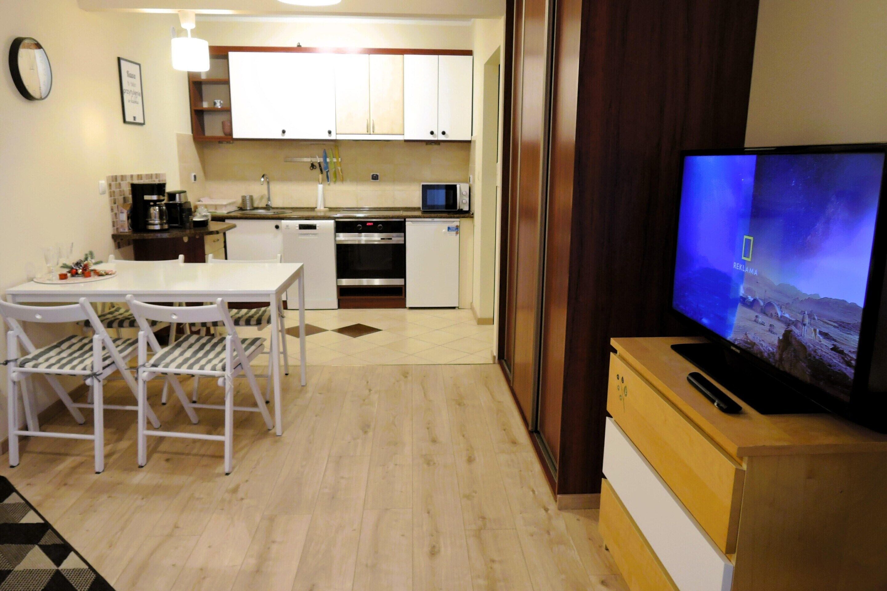 Een compacte keuken met witte kasten, een magnetron en een koffiezetapparaat. Een eettafel met stoelen staat op de voorgrond.