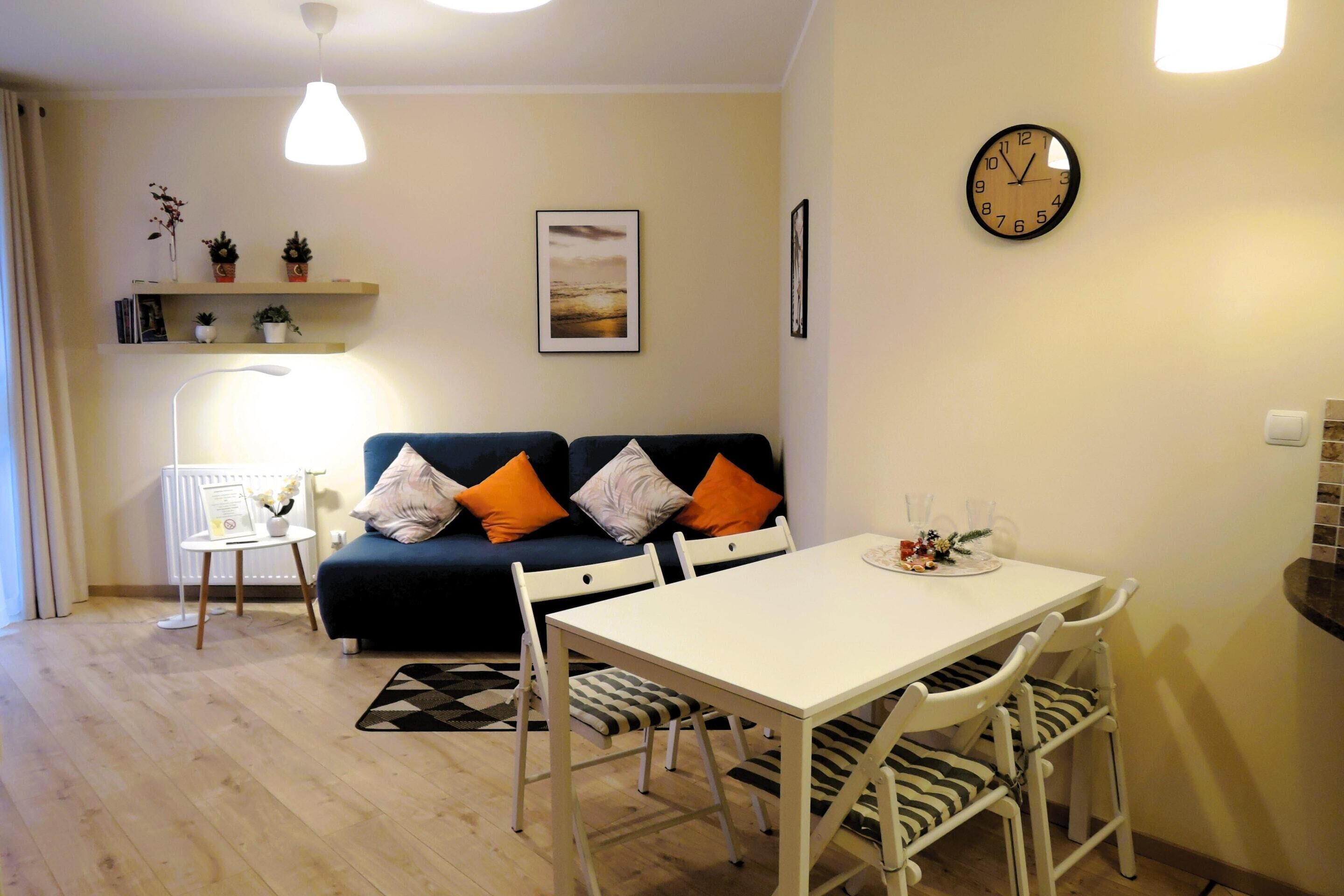 A modern living room with a white dining table, white chairs, a blue sofa with orange cushions, a small side table with a lamp, and a wall clock.