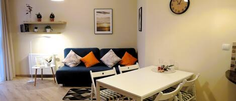 A modern living room with a white dining table, white chairs, a blue sofa with orange cushions, a small side table with a lamp, and a wall clock.