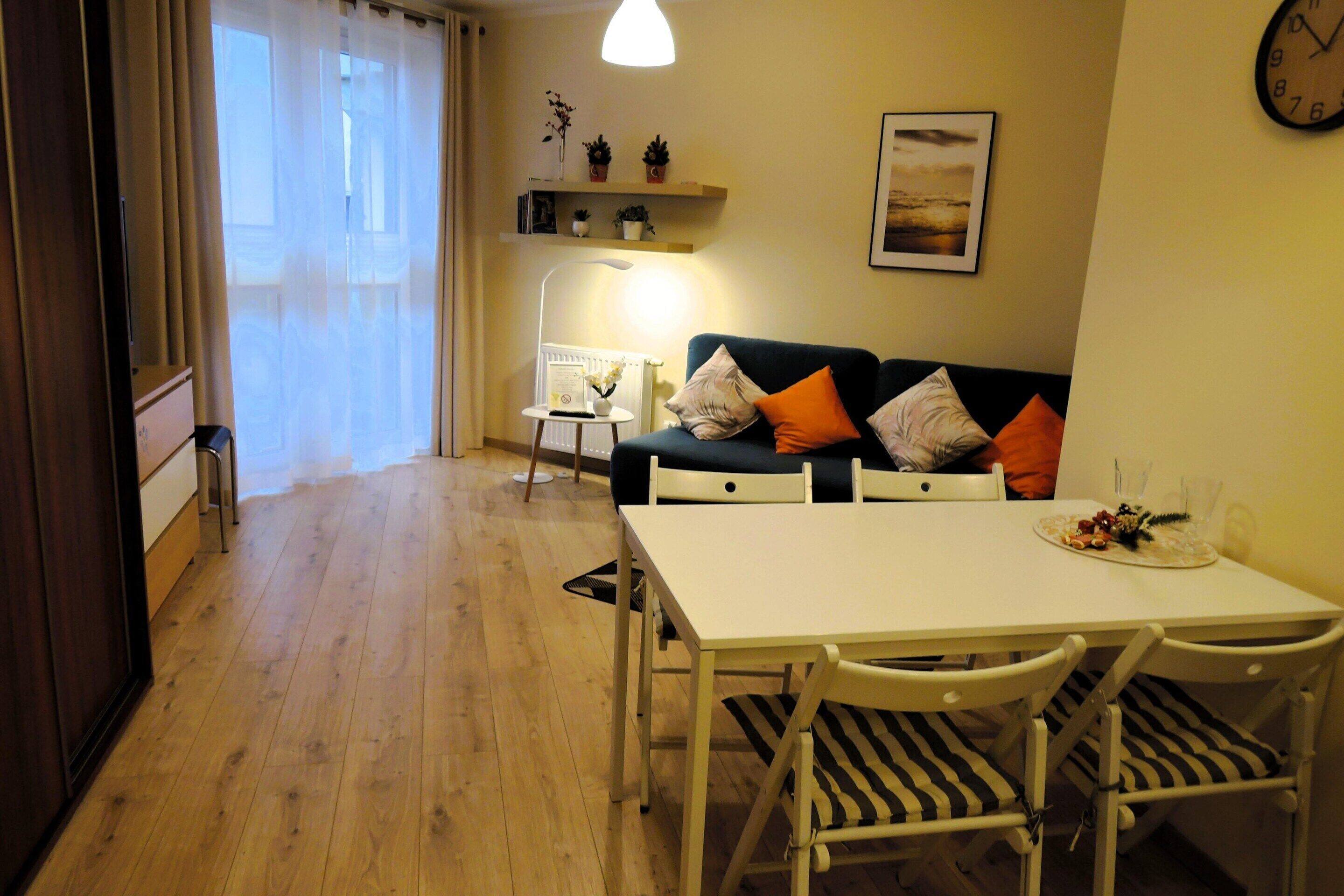 Ett mysigt vardagsrum med en soffa, ett matbord och stolar. Det står en lampa på ett sidobord, en klocka hänger på väggen och det finns en hylla med dekorativa föremål.