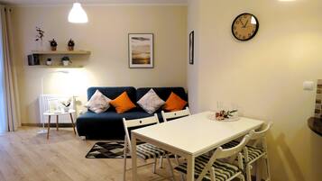 Ein modernes Wohnzimmer mit einem weißen Esstisch, weißen Stühlen, einem blauen Sofa mit orangefarbenen Kissen, einer Stehlampe und einer Wanduhr.