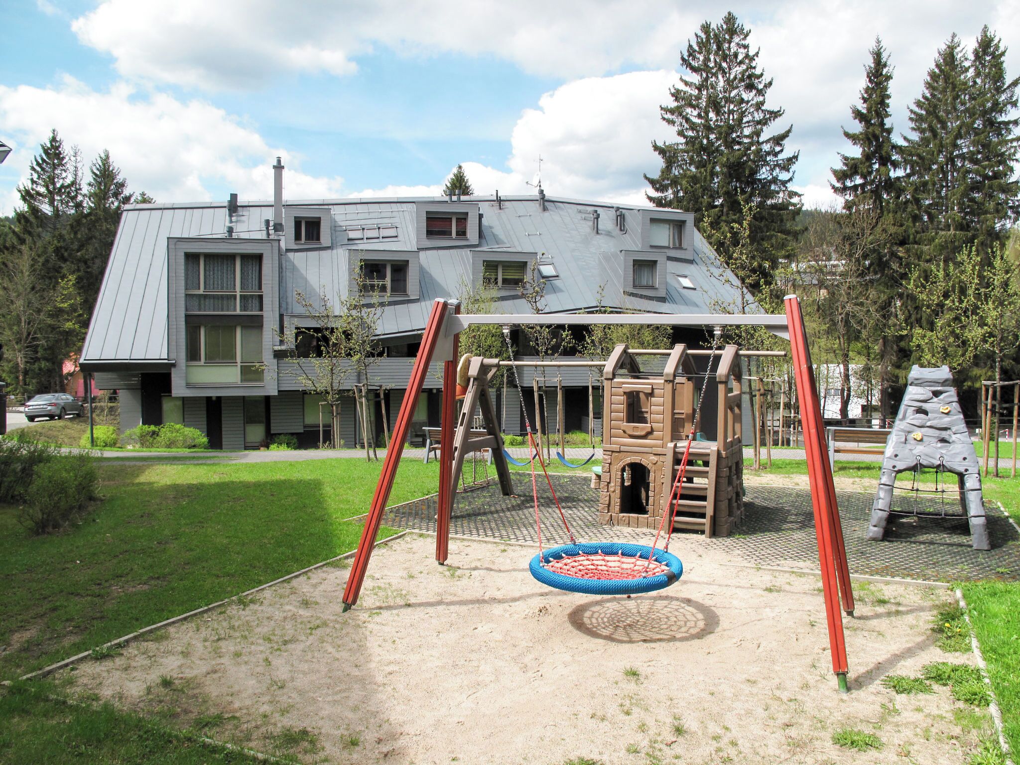 A playground with swings and a climbing structure, a building with a sloped roof in the background, and a grassy area with trees.