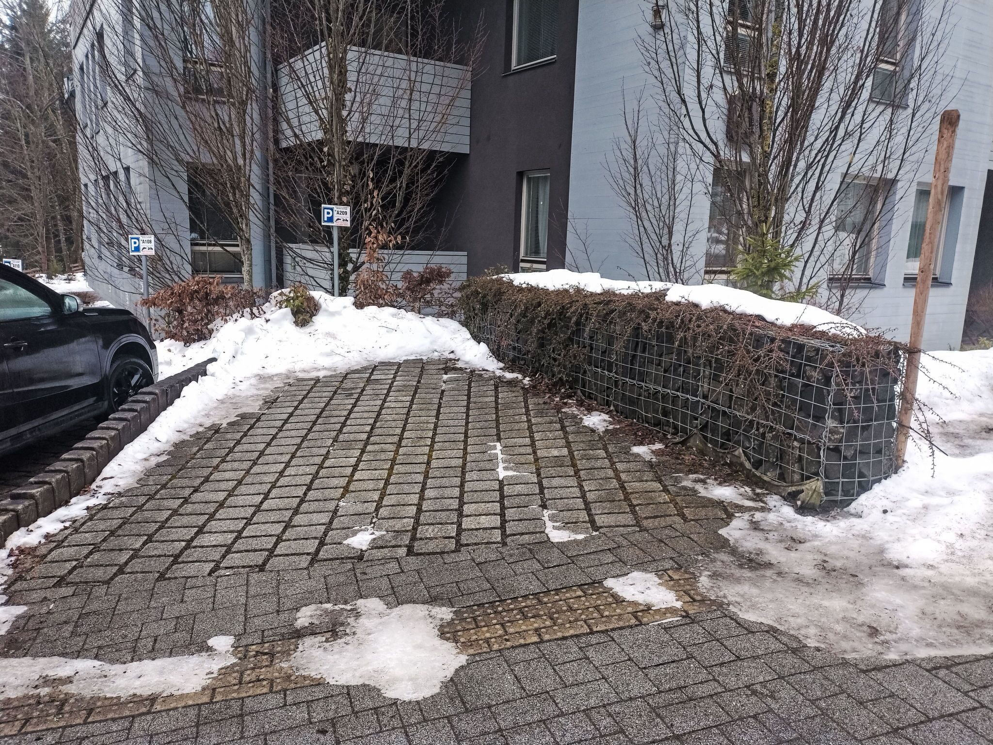 A paved pathway with snow patches, a black car parked on the left, and a building with a grey facade in the background.