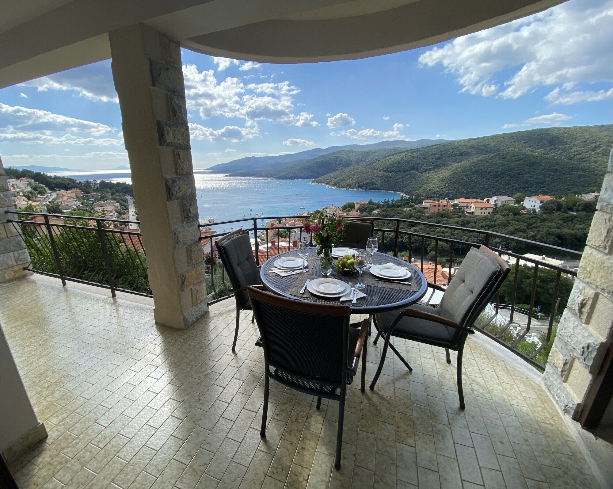 A balcony with a table set for two, overlooking a coastal landscape with buildings and hills.
