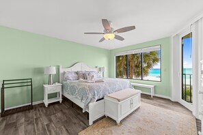A bedroom with a bed, nightstands, a bench, a ceiling fan, and a large window with a view of the beach. - Beachfront escape - steps from the Gulf (Panama City Beach)