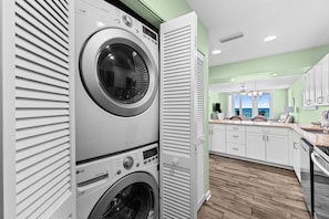 A laundry room with stacked washers and dryers, a kitchen with white cabinets, and a view through an open door. - Beachfront escape - steps from the Gulf (Panama City Beach)