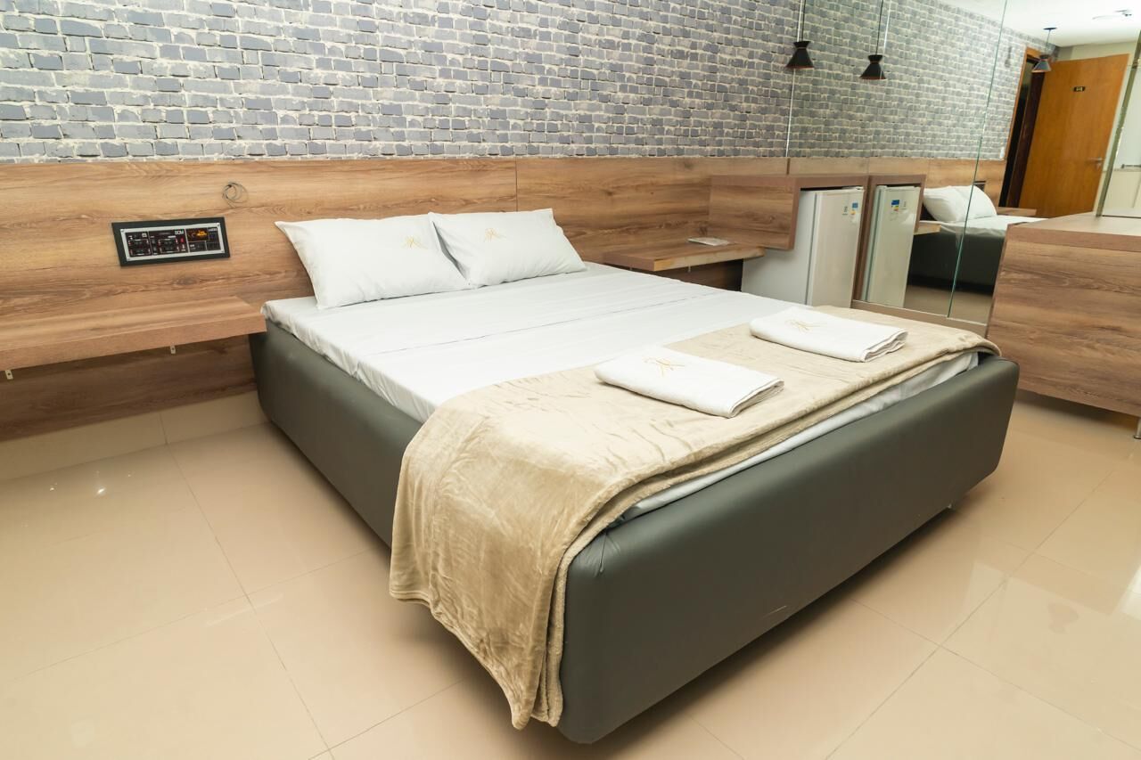 Quarto de hotel moderno com parede de tijolos, cabeceira de madeira, cama com manta bege e mesa de cabeceira com espelho.