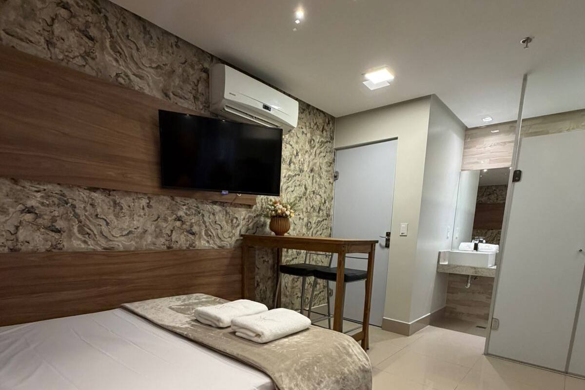 A modern bedroom with a large bed, a flat-screen TV mounted on the wall, a small table with a vase, and a bathroom with a sink and mirror.