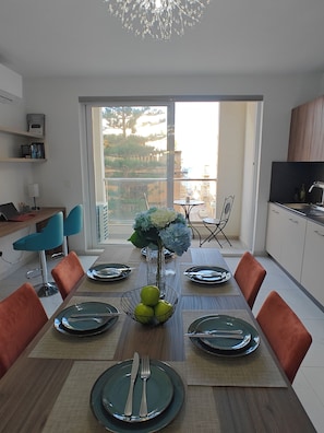 A dining area with a table set for six, a vase with flowers, and a view of the outdoors through a large window. - The Exchange - Designer apartment in St Paul's Bay with views / 300 m from beach (St Paul's Bay)