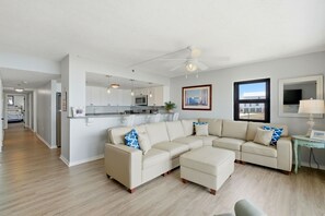 A spacious living area with a large L-shaped sectional sofa, a small table, and a ceiling fan. A kitchen is visible in the background with modern appliances and pendant lighting. - Ocean Breesze West 201 - New Listing!! Spacious Gulf Front Property! (Perdido Key)