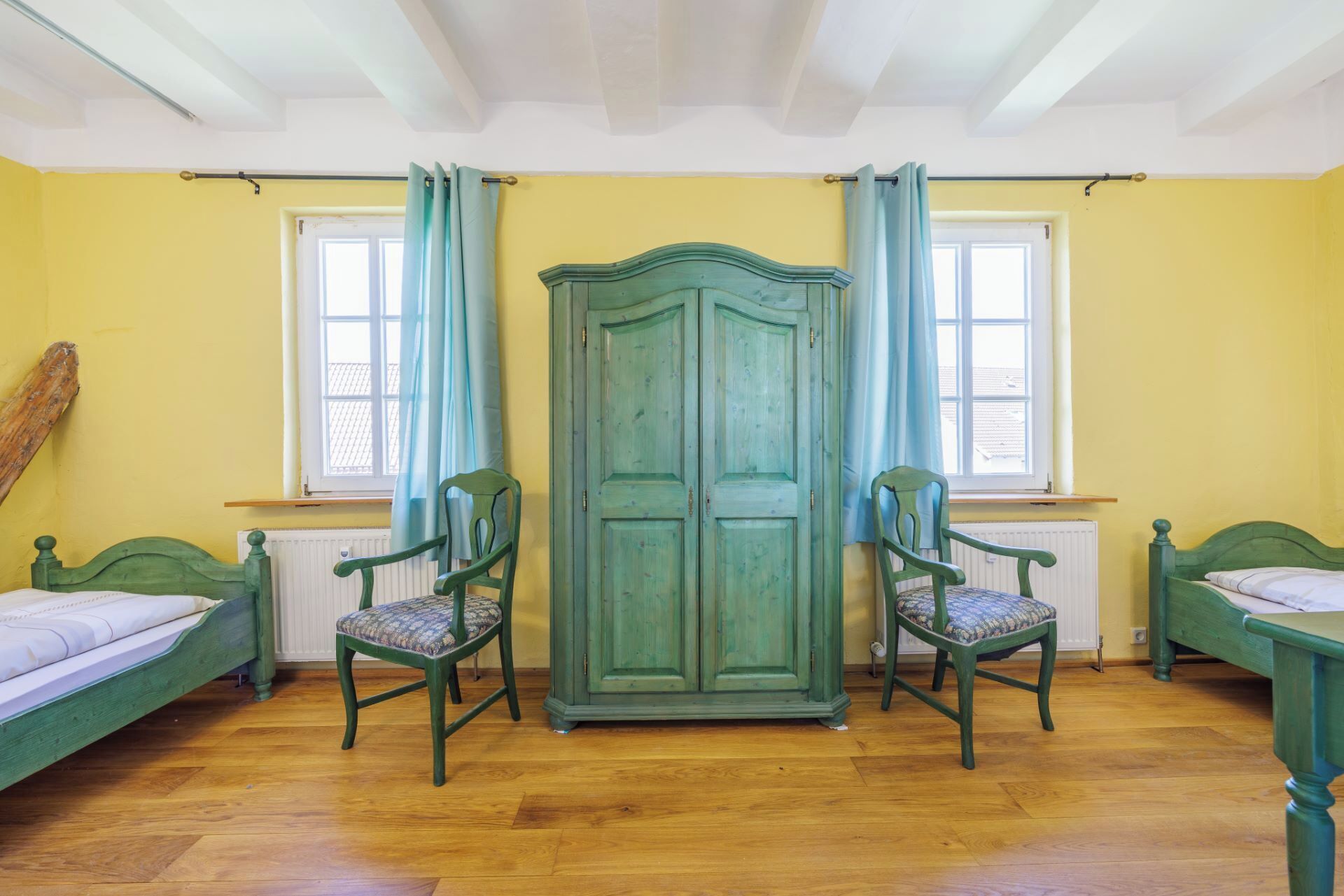 A room with two green beds, a large green wardrobe, two chairs, and two windows with curtains.