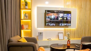 A modern living room with a flat-screen TV mounted on the wall displaying multiple channels, a sofa, a coffee table, and a bookshelf with decorative items. - Cozy 2BED w/ Private Cinema @ Bodija, Ibadan (Ibadan)