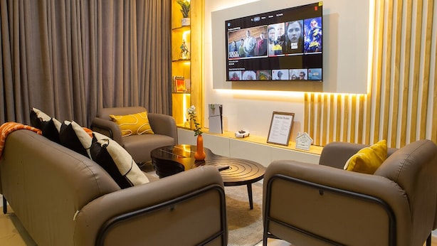 A modern living room with a flat-screen TV mounted on the wall displaying various content. There are two sofas with cushions, a coffee table, and a small side table with a vase. - Cozy 2BED w/ Private Cinema @ Bodija, Ibadan (Ibadan)