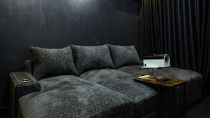 A dark-themed room with a sectional sofa, a side table, and a projector on a wooden stand. - Cozy 2BED w/ Private Cinema @ Bodija, Ibadan (Ibadan)