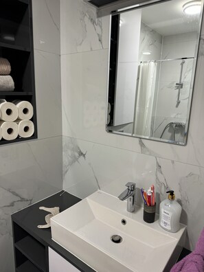 Ein Badezimmer mit einem weißen Waschbecken, einem Spiegel und einem Ablagebereich mit Toilettenpapier und Pflegeprodukten.