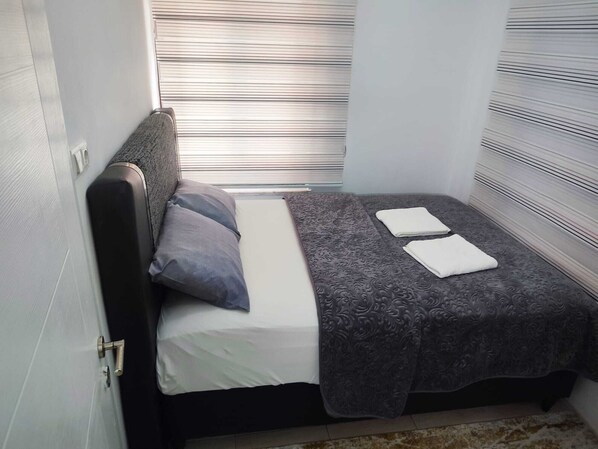 A small, neatly made bed with a grey blanket and pillows, a window with blinds, and a door with a handle.