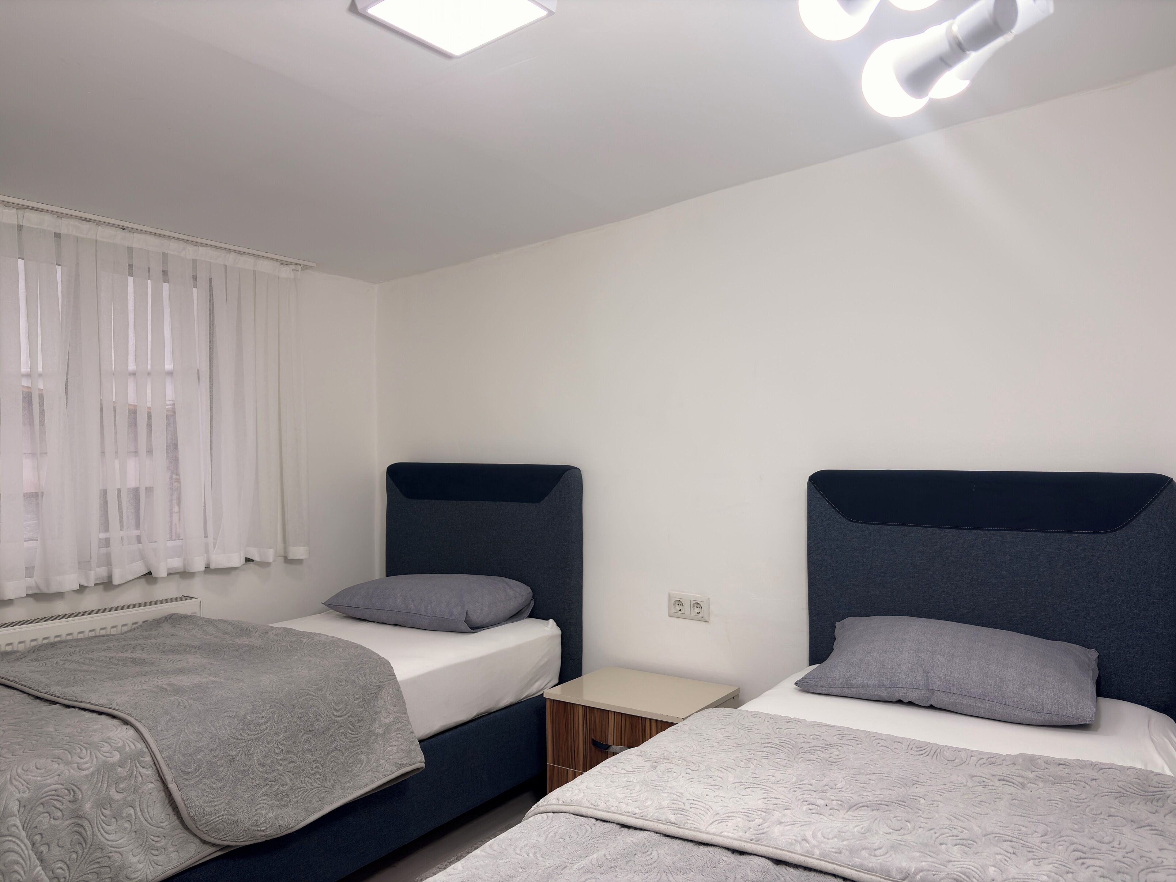 Un dormitorio moderno con dos camas, una mesita de noche y un ventanal con cortinas translúcidas.