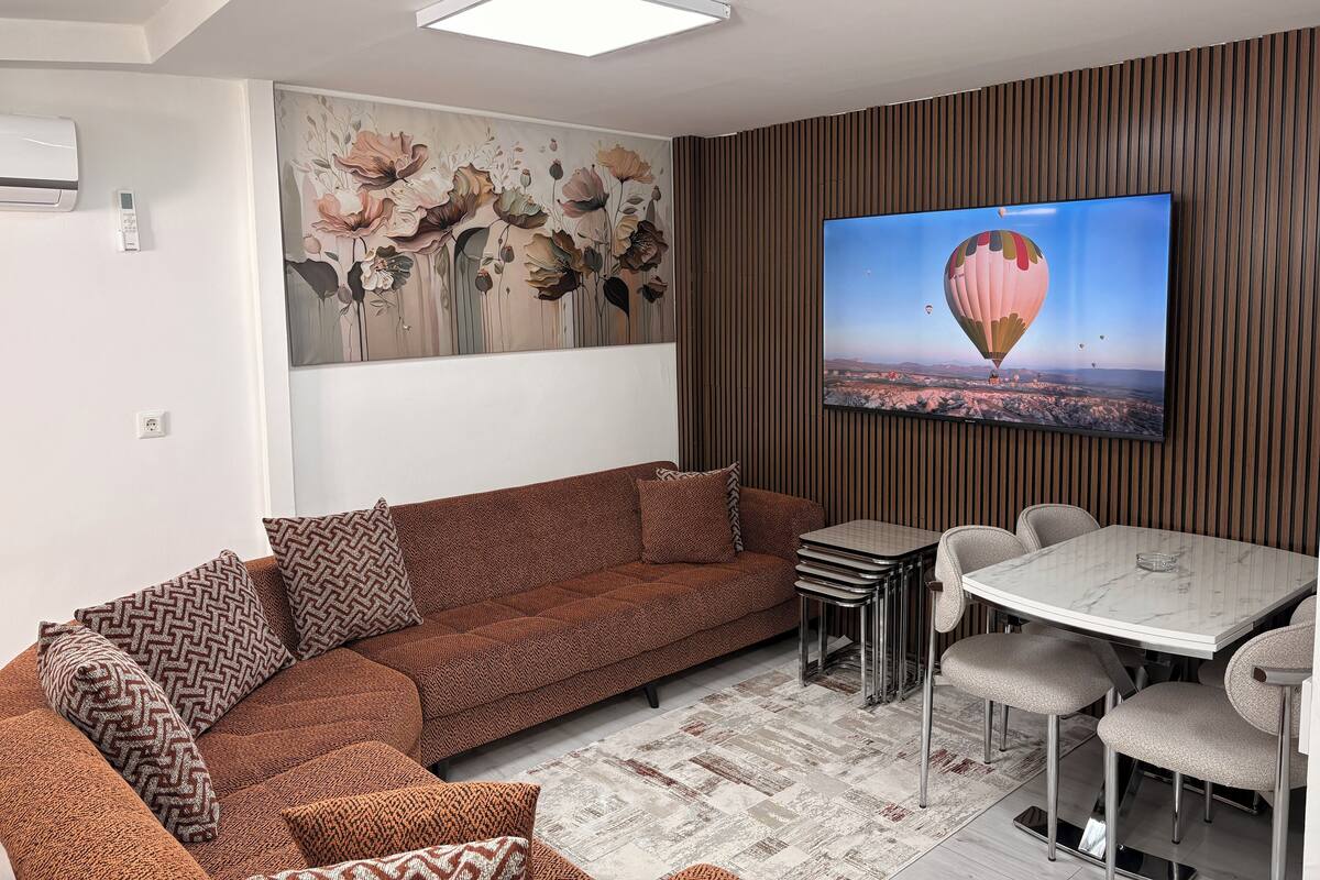 A modern living room with a sectional sofa, a dining table with chairs, a large wall-mounted TV, and a decorative floral painting.