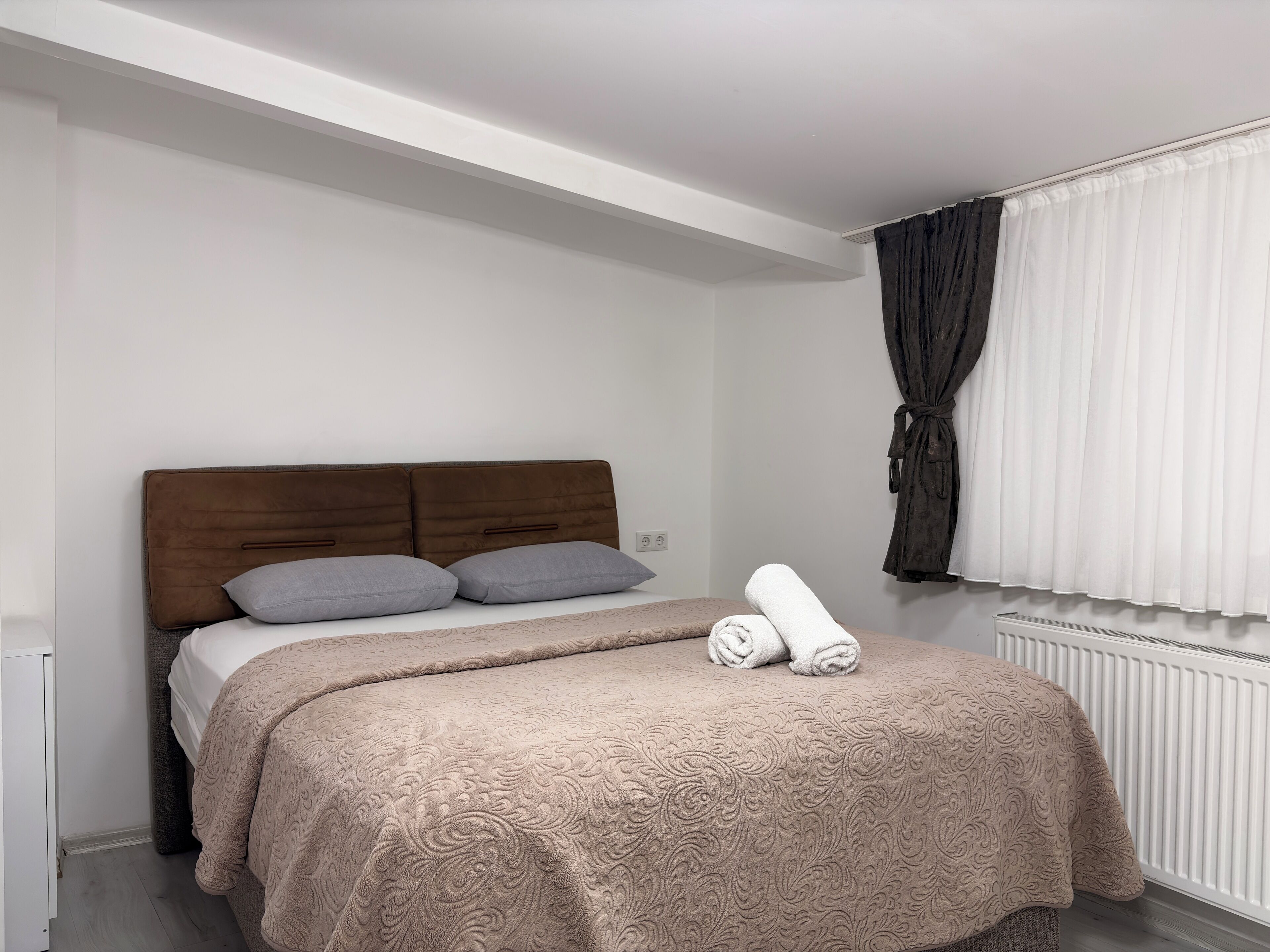 A bedroom with a bed, pillows, a rolled towel, a window with curtains, and a radiator.